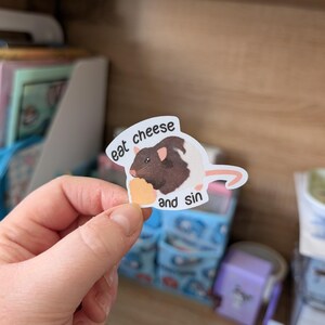 Op de afbeelding: Een sticker met een cartoonrat met bruin en wit haar, die een stuk kaas vasthoudt. De sticker heeft de tekst "eat cheese and sin" in een witte tekstballon. De achtergrond is wazig.