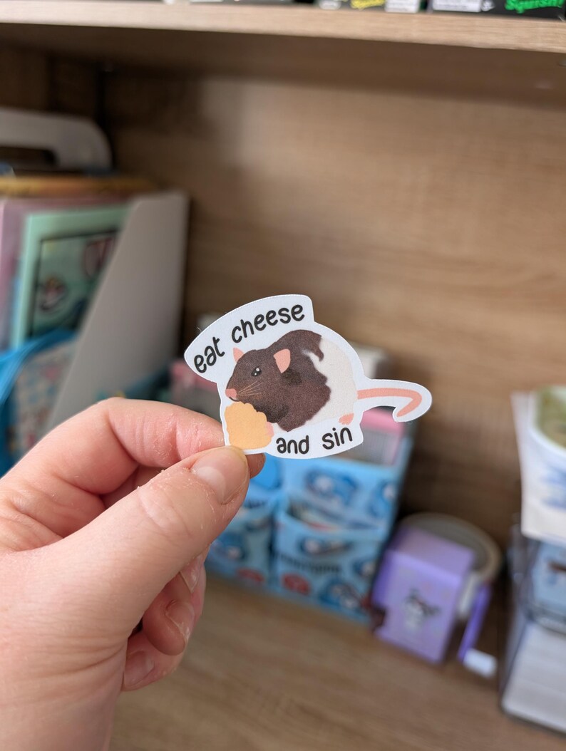 Op de afbeelding: Een sticker met een cartoonrat met bruin en wit haar, die een stuk kaas vasthoudt. De sticker heeft de tekst "eat cheese and sin" in een witte tekstballon. De achtergrond is wazig.