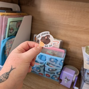 Op de afbeelding: Een sticker met een cartoon illustratie van een bruine cavia die kaas eet. De sticker heeft de tekst "eat cheese and sin" erop gedrukt. De sticker wordt in een hand gehouden.
