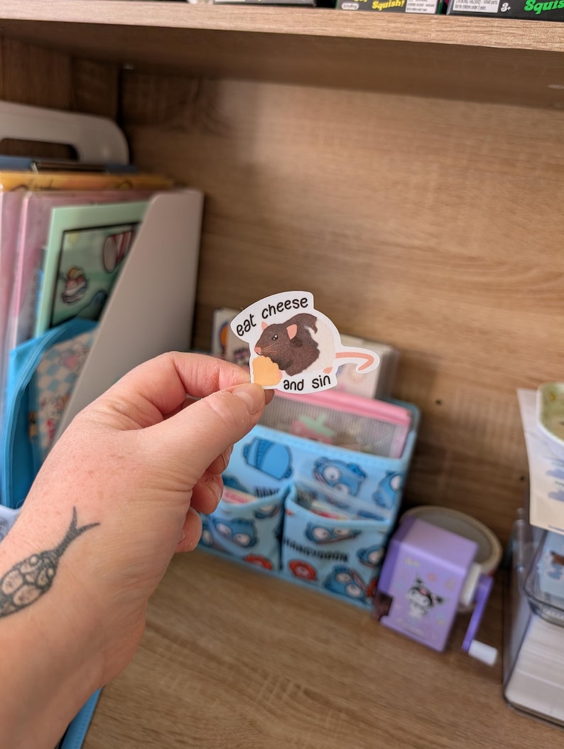 Op de afbeelding: Een sticker met een cartoon illustratie van een bruine cavia die kaas eet. De sticker heeft de tekst "eat cheese and sin" erop gedrukt. De sticker wordt in een hand gehouden.