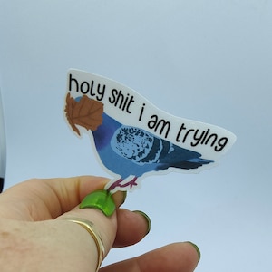 May include: A sticker depicting a blue pigeon with a brown leaf, and the text "holy shit i am trying". The sticker is held by a hand with green nail polish. The background is plain white.