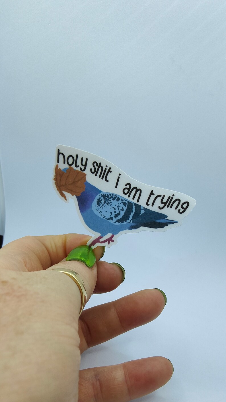 May include: A sticker depicting a blue pigeon with a brown leaf, and the text "holy shit i am trying". The sticker is held by a hand with green nail polish. The background is plain white.