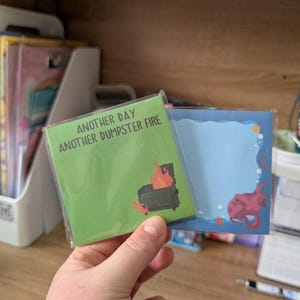 May include: Two square sticky note pads in clear plastic sleeves. The green pad features a black dumpster on fire with the text "ANOTHER DAY ANOTHER DUMPSTER FIRE". The blue pad has a scalloped border with a pink octopus and small orange stars.