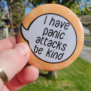 May include: A round orange button with a white speech bubble that says "I have panic attacks be kind".