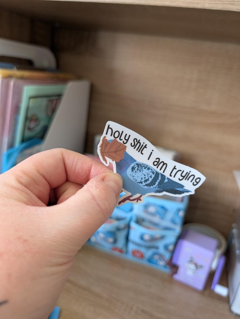 May include: A sticker depicting a blue pigeon with a brown leaf and the text "holy shit i am trying". The sticker is held up against a background of a wooden shelf and blue patterned objects.