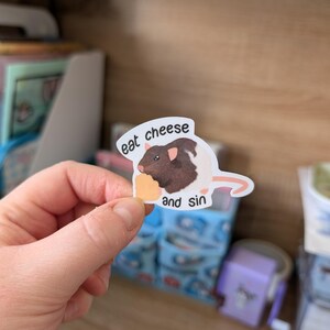 Op de afbeelding: Een sticker met een cartoonrat met bruine en witte vacht, die een stuk kaas vasthoudt. De sticker heeft de tekst "eat cheese and sin". De sticker wordt in een hand gehouden.