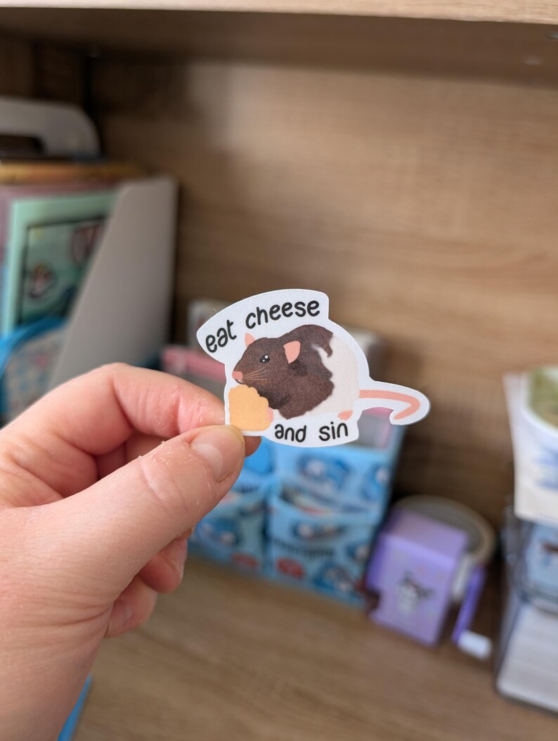 Op de afbeelding: Een sticker met een cartoonrat met bruine en witte vacht, die een stuk kaas vasthoudt. De sticker heeft de tekst "eat cheese and sin". De sticker wordt in een hand gehouden.