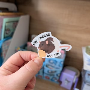 Op de afbeelding: Een sticker met een cartoonrat die kaas eet, met de tekst "eat cheese and sin". De rat is bruin en wit en de kaas is geel. De sticker heeft een witte rand.