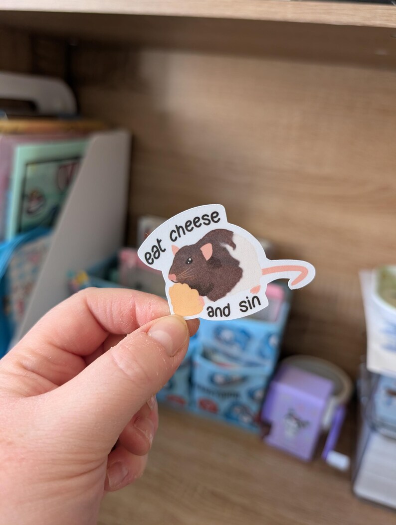 Op de afbeelding: Een sticker met een cartoonrat die kaas eet, met de tekst "eat cheese and sin". De rat is bruin en wit en de kaas is geel. De sticker heeft een witte rand.