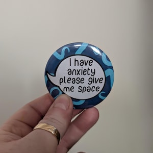 May include: A blue and white button with a speech bubble that says "I have anxiety please give me space".