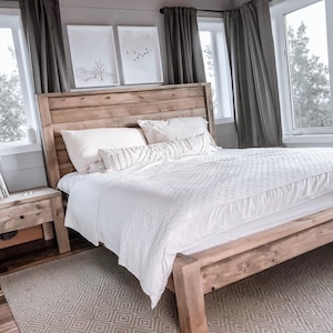 May include: A wooden bed frame with a high headboard, covered with a white duvet and pillows. The bedroom has a neutral colour scheme, with a patterned rug, grey curtains, and framed artwork. A wooden bedside table with a drawer is next to the bed.