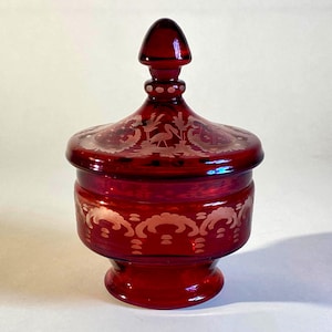Puede incluir: Un cuenco de cristal rojo intenso con tapa a juego que presenta un remate puntiagudo. El cristal está decorado con patrones blancos grabados, incluyendo una figura de ave central en la tapa y un diseño festoneado repetido en el cuerpo.