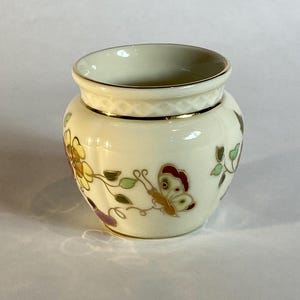 Może przedstawiać: Mały, kremowy porcelanowy pojemnik z karbowanym korpusem i złoconym brzegiem. Zewnętrzna strona ozdobiona jest delikatnym malowanym wzorem przedstawiającym kolorowe kwiaty, zielone liście oraz motyla z czerwonymi i żółtymi skrzydłami, z motywem rombów.