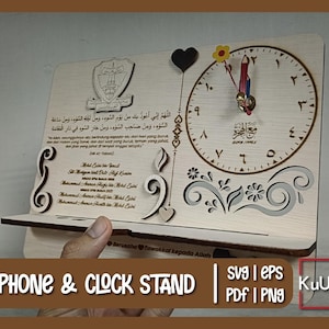 May include: A wooden phone and clock stand with a decorative clock face, floral design, and Arabic text. The stand is made of light wood and has a cutout design. The clock face is white with black numbers and hands. The floral design is in a light gray color. The Arabic text is in black. The stand is labeled "Phone & Clock Stand" and "KuUkir".