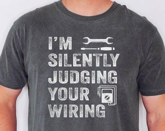 May include: Dark gray t-shirt with the text "I'M SILENTLY JUDGING YOUR WIRING" in white. The design includes a wrench, screwdriver, and a multimeter. The shirt is a crew neck and has short sleeves.