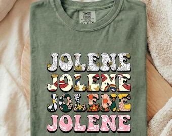 May include: An olive green t-shirt featuring the name JOLENE printed four times in a stacked block format. Each line of text displays a unique pattern, including white daisies, red lips with rainbows, orange and yellow flowers, and pink floral designs.