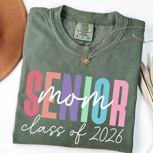 May include: Olive green t-shirt with the words "SENIOR mom class of 2026" in colorful block letters and script. A gold necklace with a pendant is draped across the shirt. A brown hat is in the upper left corner.