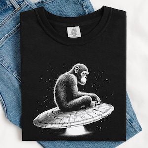 May include: Black t-shirt featuring a detailed black and white illustration of a chimpanzee seated on a UFO. The design includes a starry background. The shirt is made by Comfort Colors.