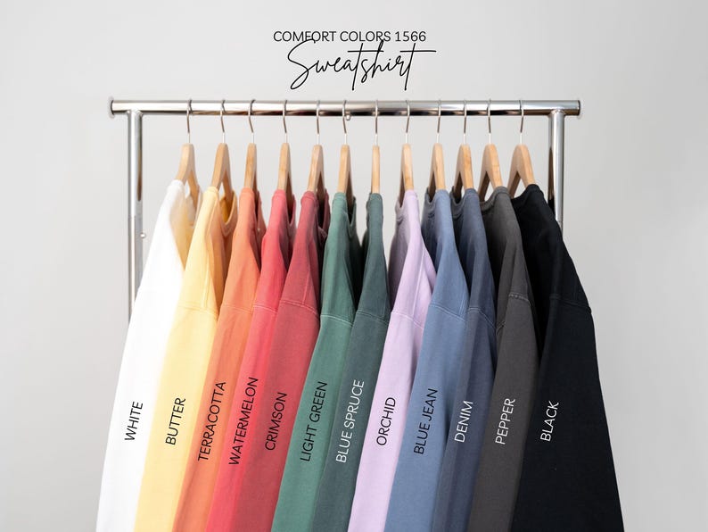 May include: A clothing rack displays a variety of Comfort Colors 1566 sweatshirts in various colors, including white, yellow, red, green, blue, and black. The text "Sweatshirt" is written in a script font above the shirts.