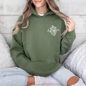 May include: Olive green hoodie with a white anatomical heart and heartbeat line graphic on the front. The hoodie has a drawstring hood and a kangaroo pocket. The model is wearing light wash jeans.