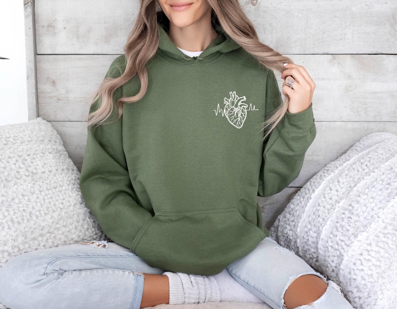 May include: Olive green hoodie with a white anatomical heart and heartbeat line graphic on the front. The hoodie has a drawstring hood and a kangaroo pocket. The model is wearing light wash jeans.
