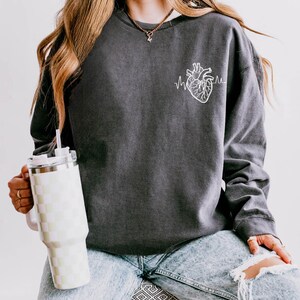 May include: Dark gray sweatshirt featuring a white anatomical heart graphic with a heartbeat line. The sweatshirt is worn with light wash ripped jeans. A checkered tumbler with a straw is held in the hand.