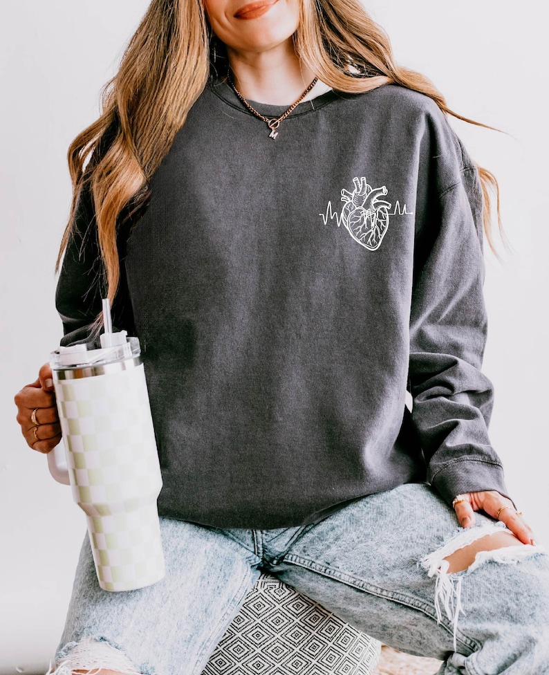 May include: Dark gray sweatshirt featuring a white anatomical heart graphic with a heartbeat line. The sweatshirt is worn with light wash ripped jeans. A checkered tumbler with a straw is held in the hand.
