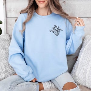 May include: A light blue sweatshirt featuring a black anatomical heart design with a heartbeat line. The sweatshirt is a solid light blue color and has a crew neck. The design is on the upper left chest area.