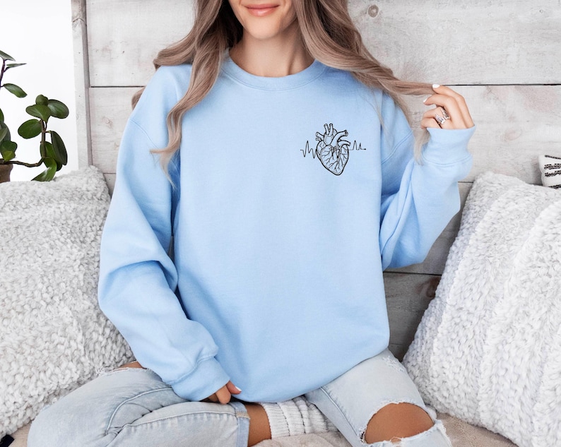 May include: A light blue sweatshirt featuring a black anatomical heart design with a heartbeat line. The sweatshirt is a solid light blue color and has a crew neck. The design is on the upper left chest area.