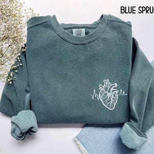 May include: Blue spruce-colored sweatshirt with a white anatomical heart and heartbeat line graphic. The text "BLUE SPRUCE" is at the top. The sweatshirt has a crew neck and cuffed sleeves. The brand "Comfort Colors" is on the label.