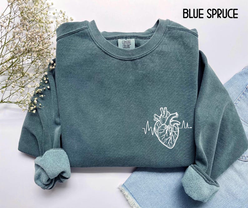 May include: Blue spruce-colored sweatshirt with a white anatomical heart and heartbeat line graphic. The text "BLUE SPRUCE" is at the top. The sweatshirt has a crew neck and cuffed sleeves. The brand "Comfort Colors" is on the label.