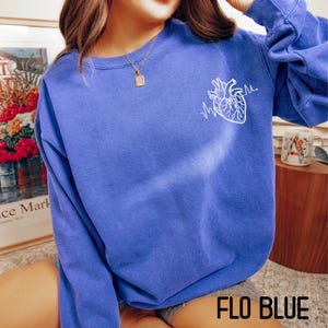 May include: A blue sweatshirt with a white anatomical heart and heartbeat line graphic. The text "FLO BLUE" is at the bottom of the image. The sweatshirt is a solid blue color.