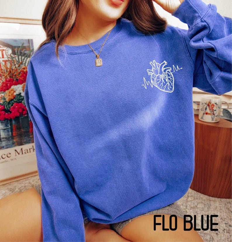May include: A blue sweatshirt with a white anatomical heart and heartbeat line graphic. The text "FLO BLUE" is at the bottom of the image. The sweatshirt is a solid blue color.