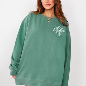 May include: A teal green sweatshirt with a white embroidered anatomical heart design. The crew neck sweatshirt is oversized and has long sleeves. The model is wearing a gold necklace. The sweatshirt is a casual, comfortable style.