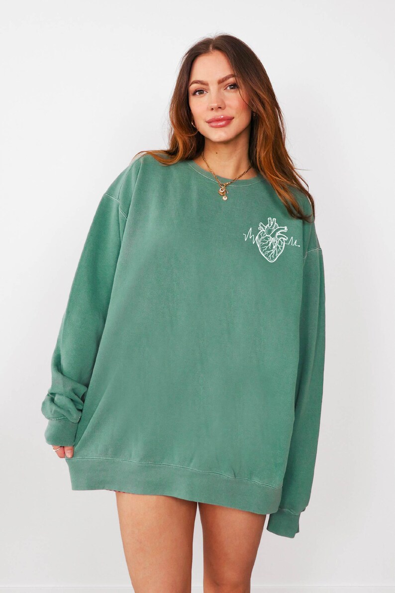 May include: A teal green sweatshirt with a white embroidered anatomical heart design. The crew neck sweatshirt is oversized and has long sleeves. The model is wearing a gold necklace. The sweatshirt is a casual, comfortable style.