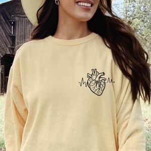 May include: A light yellow long-sleeve shirt featuring a black anatomical heart graphic with a heartbeat line. The shirt is made of a soft material.