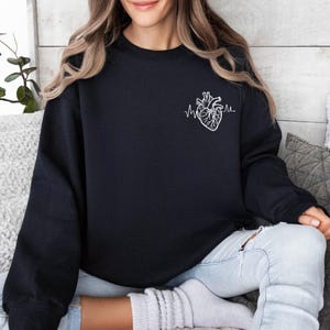 May include: Black sweatshirt featuring a white anatomical heart design with a heartbeat line. The sweatshirt is a crew neck style and is made of a soft material. The design is on the left chest area.