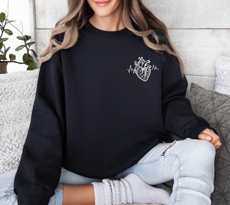 May include: Black sweatshirt featuring a white anatomical heart design with a heartbeat line. The sweatshirt is a crew neck style and is made of a soft material. The design is on the left chest area.