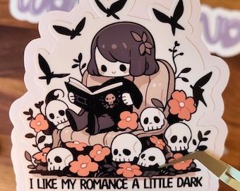 May include: A sticker featuring a cartoon illustration of a girl sitting in a chair surrounded by skulls and flowers. The girl is reading a book with a skull on the cover. The text "I like my romance a little dark" is at the bottom of the sticker.