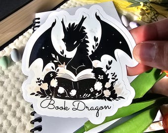 Op de afbeelding: Een zwart-witte sticker van een draak die een boek leest met de tekst "Book Dragon" eronder. De draak is omringd door bloemen en sterren.