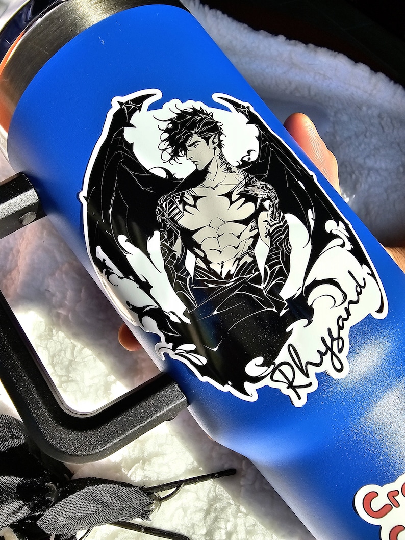 Puede incluir: Un vaso de acero inoxidable azul con una pegatina en blanco y negro de una figura musculosa alada. La pegatina tiene el nombre "Rhysand" escrito en cursiva.