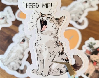 May include: A white and brown cat sticker with a white background. The cat is sitting up with its mouth open and its tongue sticking out. The sticker says "FEED ME!" in black text.