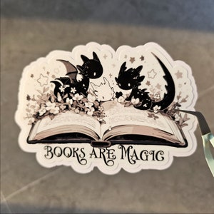 May include: A sticker featuring three black and white dragons with wings, sitting on an open book. The book has the text "Books are Magic" on the front cover.
