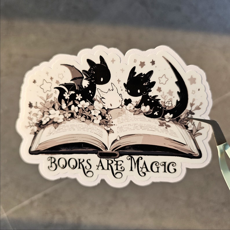May include: A sticker featuring three black and white dragons with wings, sitting on an open book. The book has the text "Books are Magic" on the front cover.