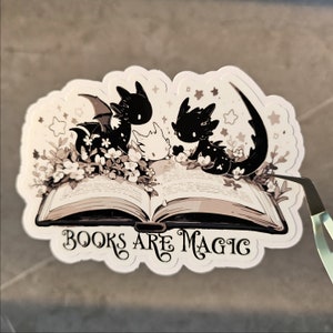 May include: A sticker featuring three black and white dragons reading a book with the text "BOOKS ARE MAGIC" on a white background.