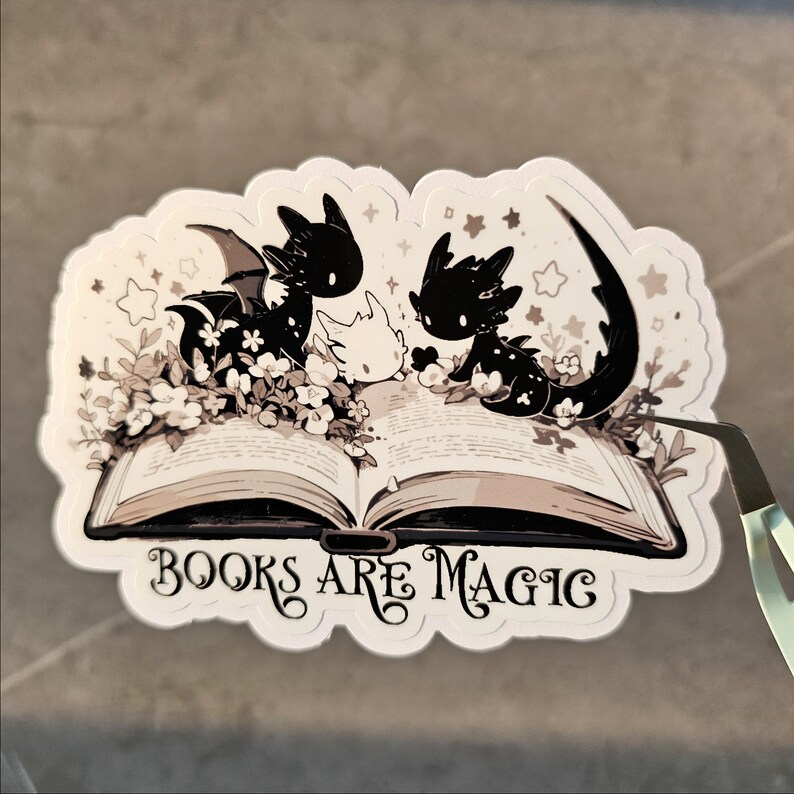 May include: A sticker featuring three black and white dragons reading a book with the text "BOOKS ARE MAGIC" on a white background.