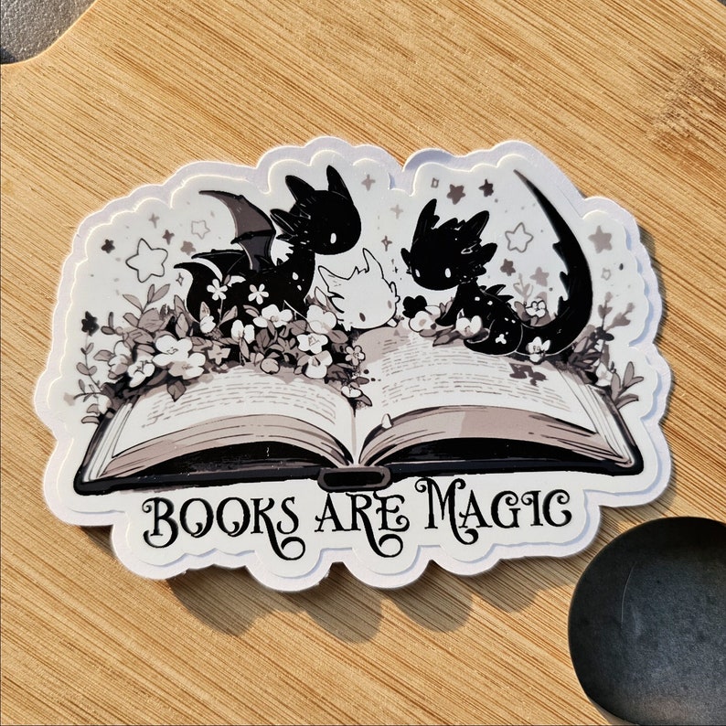 May include: A white sticker with a black and white illustration of three dragons reading a book. The book is open and the dragons are sitting on top of it. The text "BOOKS ARE MAGIC" is written below the book.