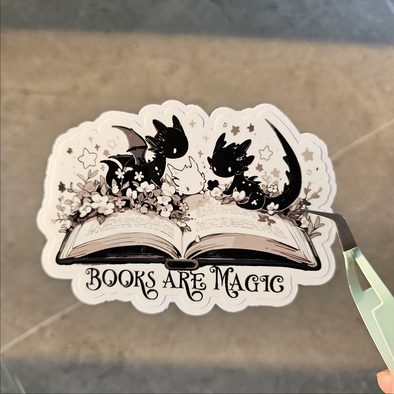 May include: A white sticker with a black and white illustration of three dragons reading a book. The book is open and the dragons are sitting on the pages. The text "BOOKS ARE MAGIC" is written below the book.
