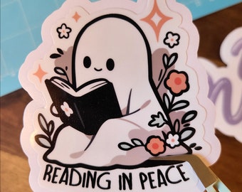 Könnte beinhalten: Ein weißer Geist mit freundlichem Gesicht liest ein schwarzes Buch, während er auf einem Bett aus rosa und weißen Blumen sitzt. Der Text "READING IN PEACE" steht unter dem Geist.