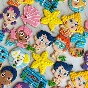 Puede incluir: Una colección de galletas de azúcar decoradas y coloridas con personajes del programa de televisión infantil, Bubble Guppies. Las galletas están decoradas con varias imágenes, incluyendo una estrella de mar, una concha, un pez y los propios personajes. Las galletas están dispuestas sobre una superficie de mármol blanco.